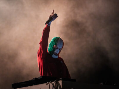 DJ Próvaí of Kneecap raising his arm on stage in an Irish tricolour balaclava during Rockwave Festival in Malakasa, Greece, 2026