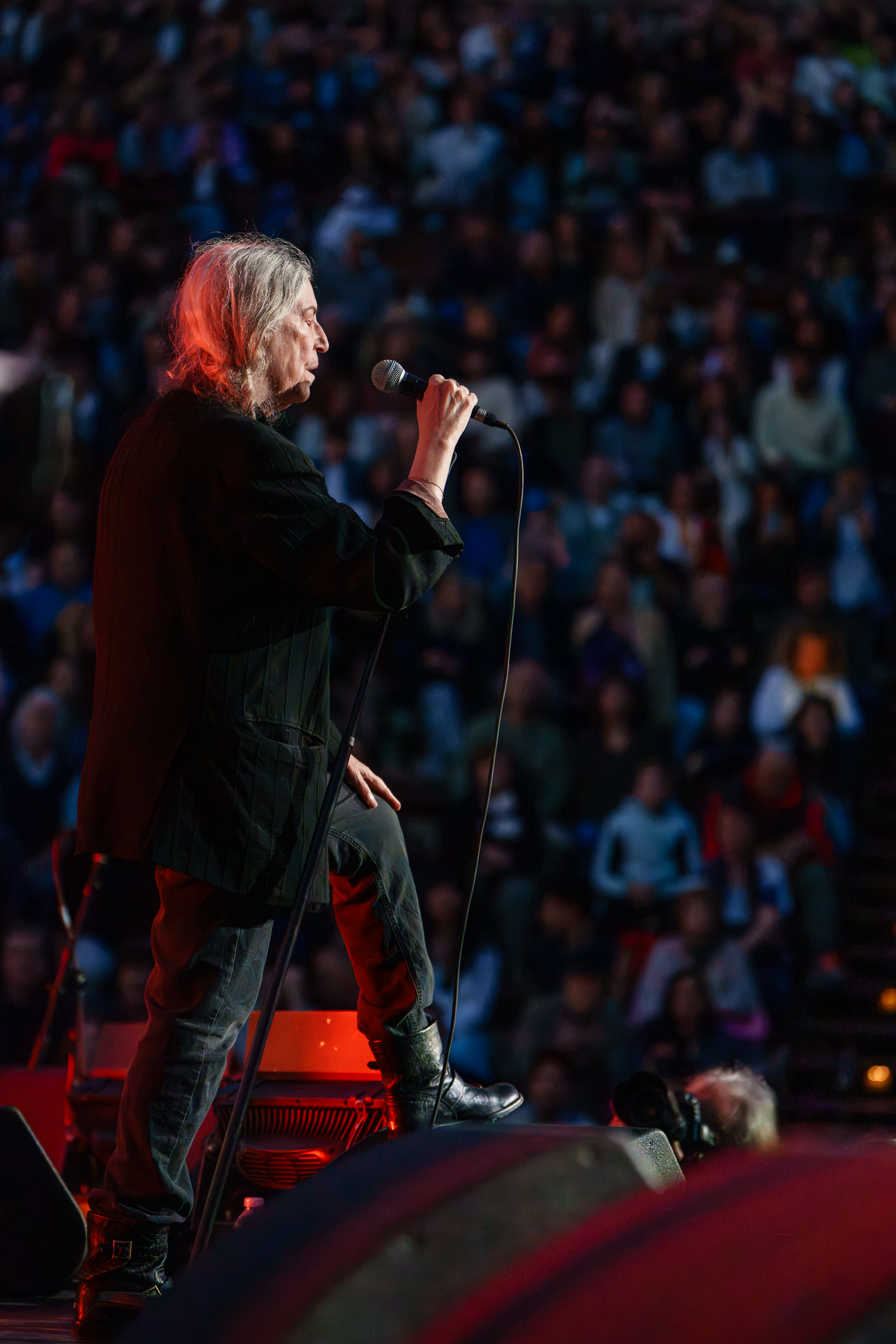 Patti Smith singing into a handheld microphone from the side of the stage, with the packed Lycabettus Theater audience filling the background